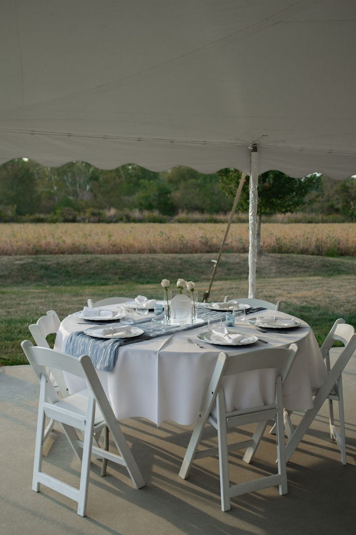 Round table with white linens, folding chairs and place settings under a white event tent
